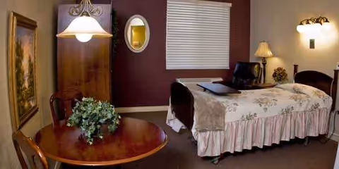 A cozy bedroom in a senior facility with a floral bedspread, armoire, table, and plants.