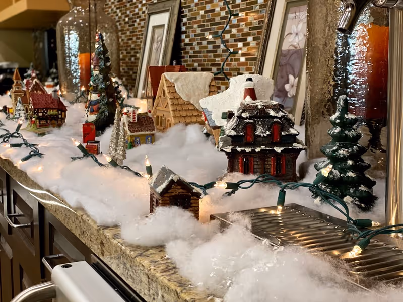 A festive holiday display on a kitchen countertop featuring miniature houses, snow-covered trees, and cotton-like snow with string lights intertwined. The background shows a mosaic tile backsplash and framed pictures.