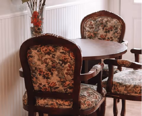 A round wooden table surrounded by four vintage-style chairs with floral upholstery in a room with white wainscoting and a vase with red flowers on the table.