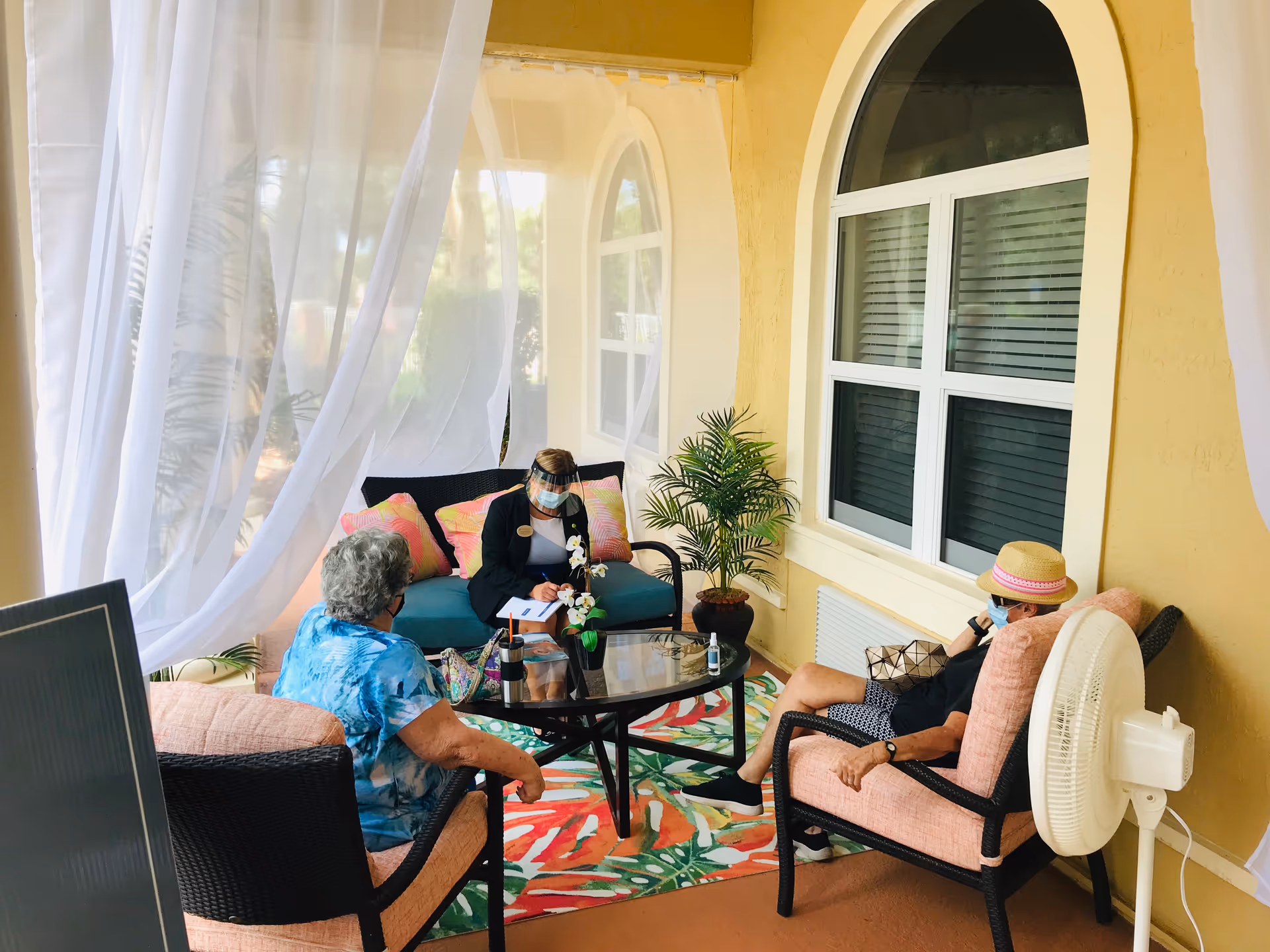 Residents and a staff member wearing masks and a face shield sit around a coffee table on a covered outdoor patio with wicker chairs, potted plants, and sheer white curtains.