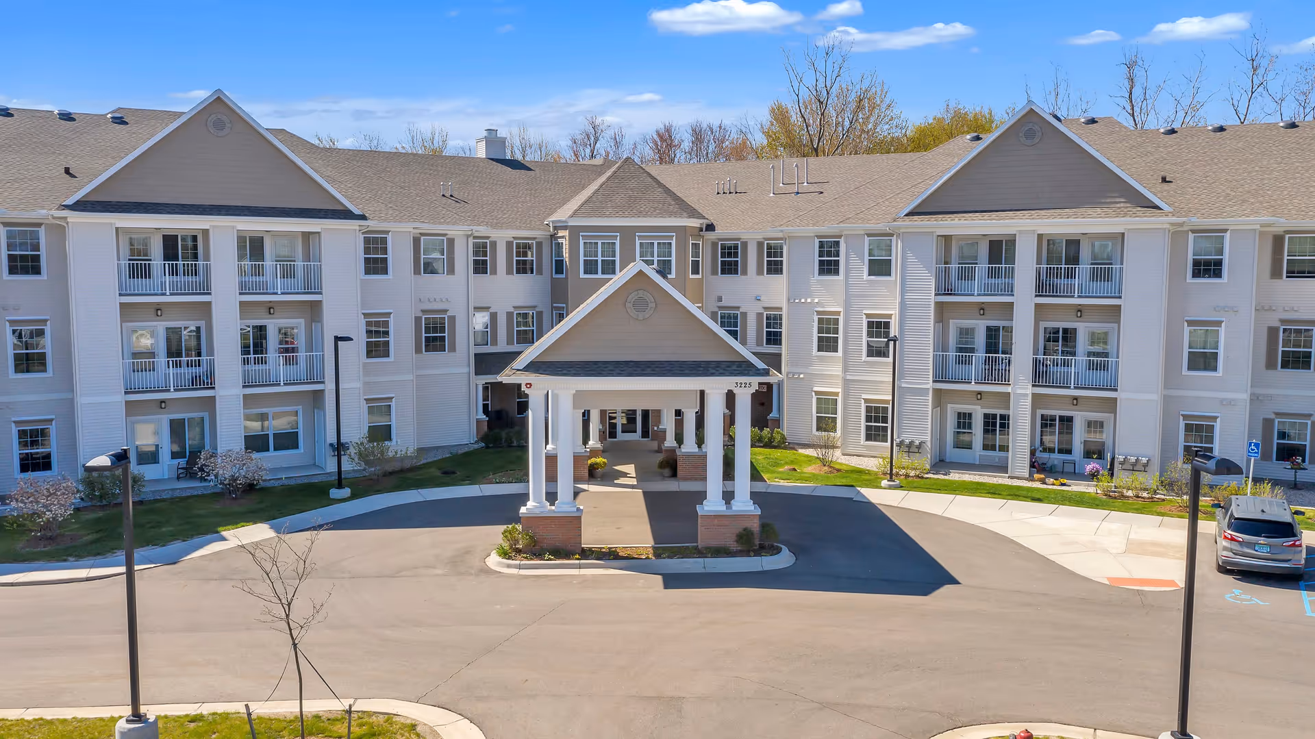 Front exterior view of a large, three-story senior living facility building with multiple balconies and a covered entrance driveway. The building is light-colored with beige and white siding, surrounded by a paved driveway and some landscaping with small trees and shrubs. A car is parked in a designated parking area to the right.