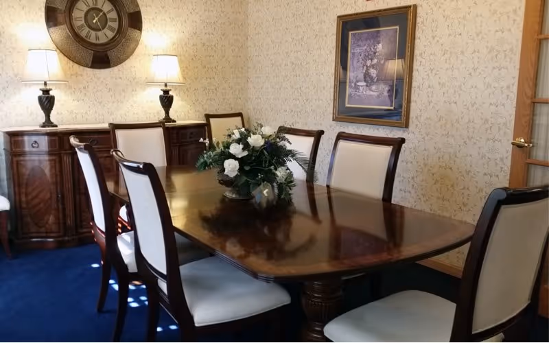 A formal dining room with a polished wooden table surrounded by six upholstered chairs. A floral centerpiece is placed on the table. The room has patterned wallpaper, a large round wall clock, two table lamps on a wooden sideboard, and a framed picture on the wall.