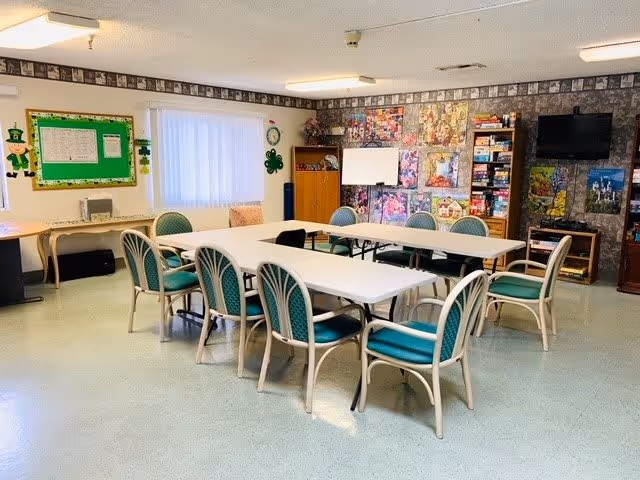 A bright activity room with two rectangular white tables arranged in a U-shape surrounded by green cushioned chairs. The room has a bulletin board with a leprechaun decoration, a window with vertical blinds, a whiteboard, shelves filled with board games, and colorful artwork on the walls. A television is mounted on the wall above a small cabinet.