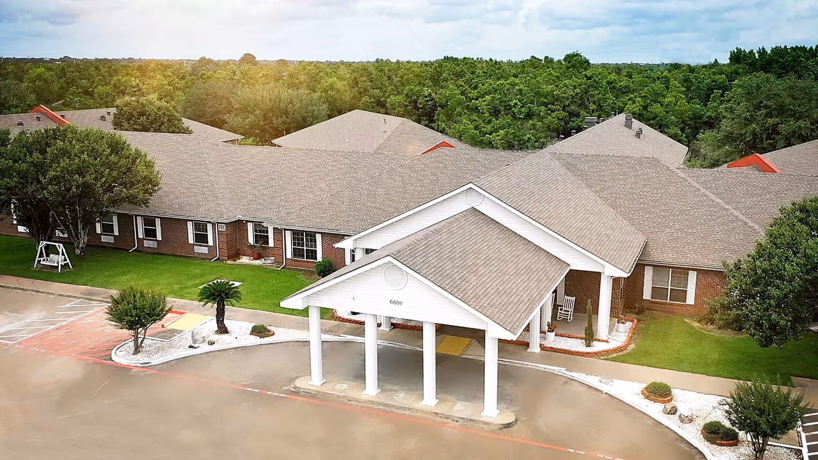 Aerial view of a single-story brick senior living facility with a white covered entrance portico, parking area, and surrounding trees.