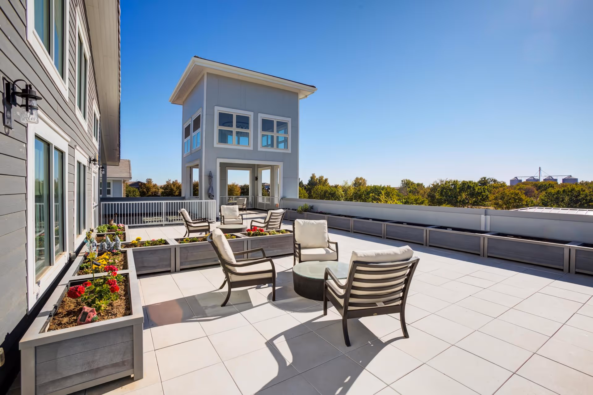 Sunlit rooftop terrace with cushioned lounge chairs, a round table, and planter boxes overlooking treetops.