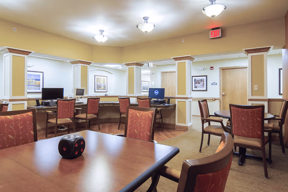 A well-lit common area in a senior living facility featuring multiple tables and chairs with red patterned upholstery. In the background, there is a curved desk area with computer workstations and framed artwork on the walls. The space has beige walls, wooden doors, and decorative columns with crown molding. Ceiling lights and an exit sign are visible.