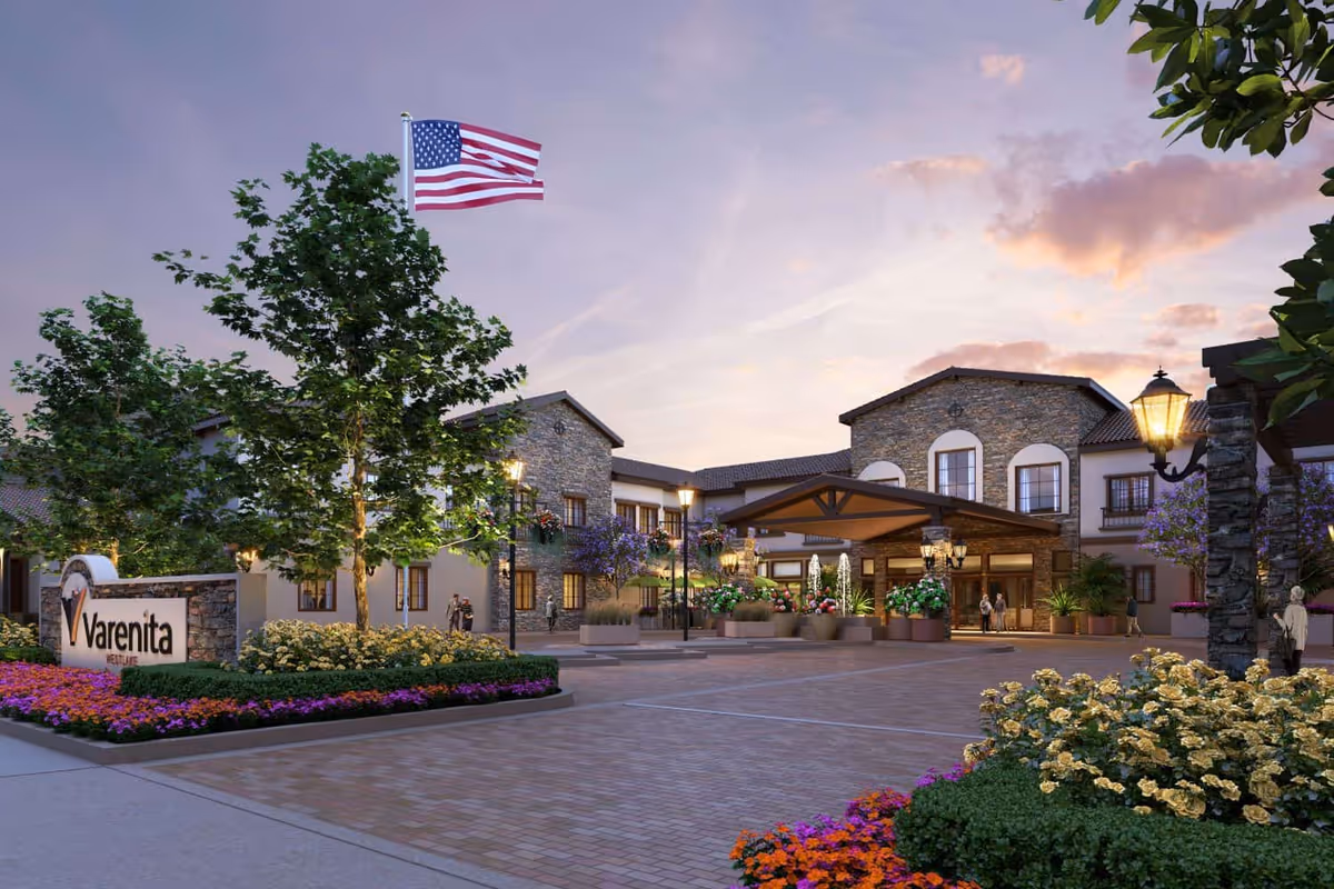 Front entrance of the Varenita senior living building with a porte-cochere, American flag, and landscaped flowerbeds at dusk.