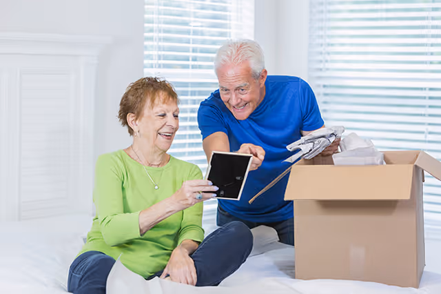 An elderly woman sitting on a bed holding a picture frame, smiling, while an elderly man beside her points at the frame and holds other items from an open cardboard box in a bright room with white blinds.
