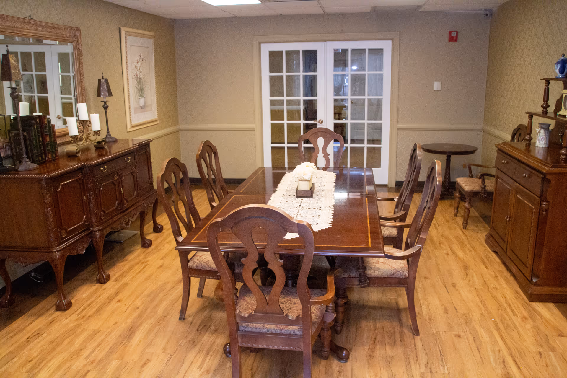 A traditional dining room with a wooden dining table and six matching chairs. The table has a lace runner and a centerpiece with white flowers. The room features wooden flooring, a large mirror above a wooden sideboard with decorative lamps and candles, framed artwork on the wall, and French doors with glass panes at the back.