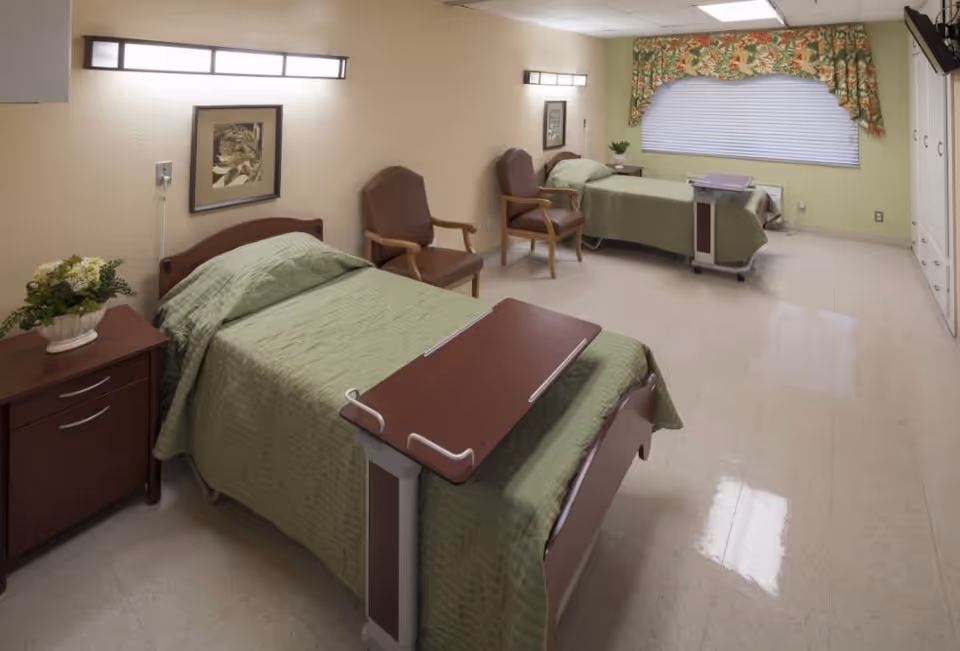 Bright shared nursing room with two hospital beds, overbed tables, chairs, and a window with floral valance.