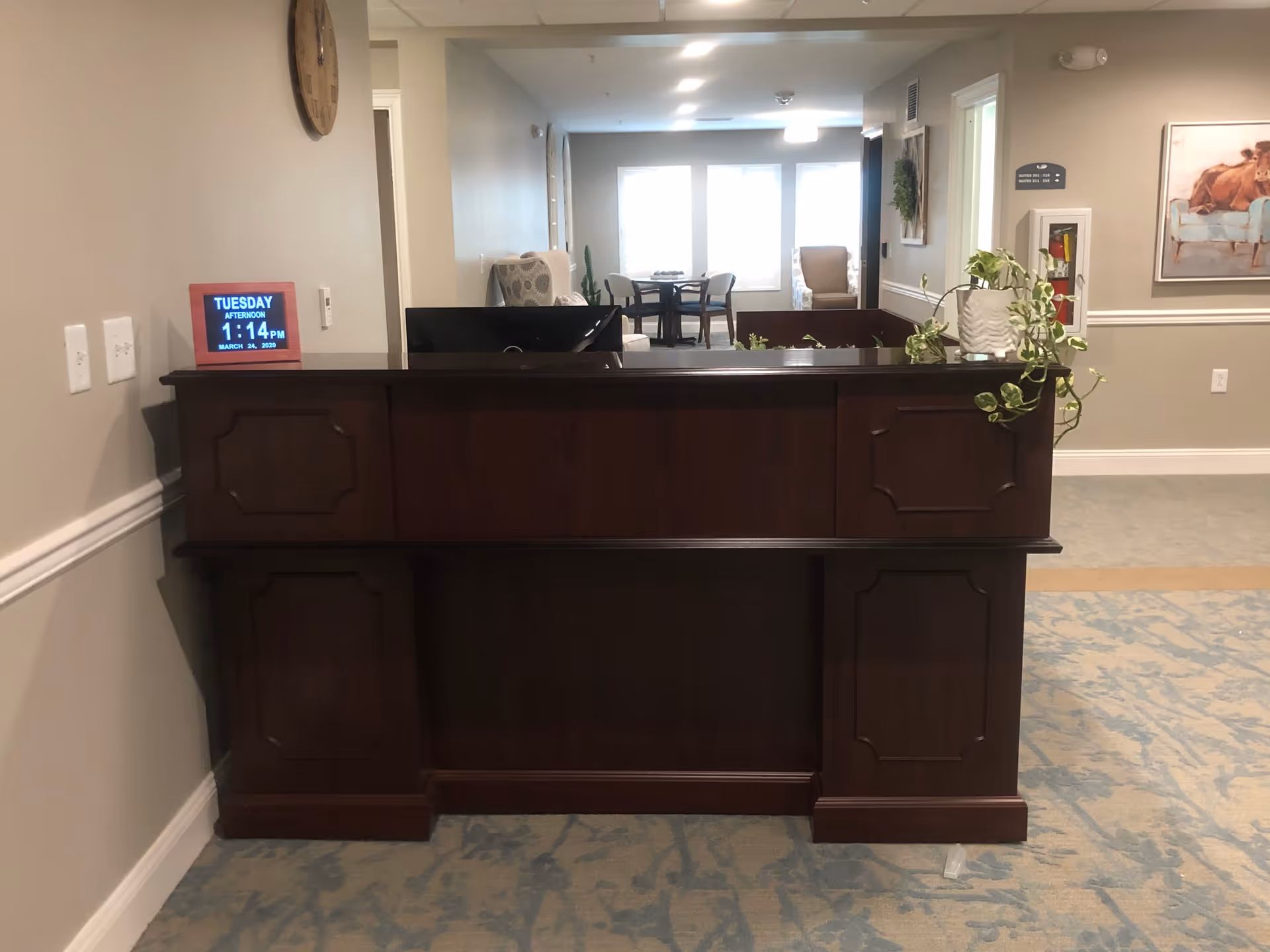 Reception desk area with a dark wooden counter, a digital clock displaying the date and time, a wall clock above, and a seating area with chairs and tables in the background. The walls are light-colored with some framed artwork and a fire extinguisher cabinet visible.