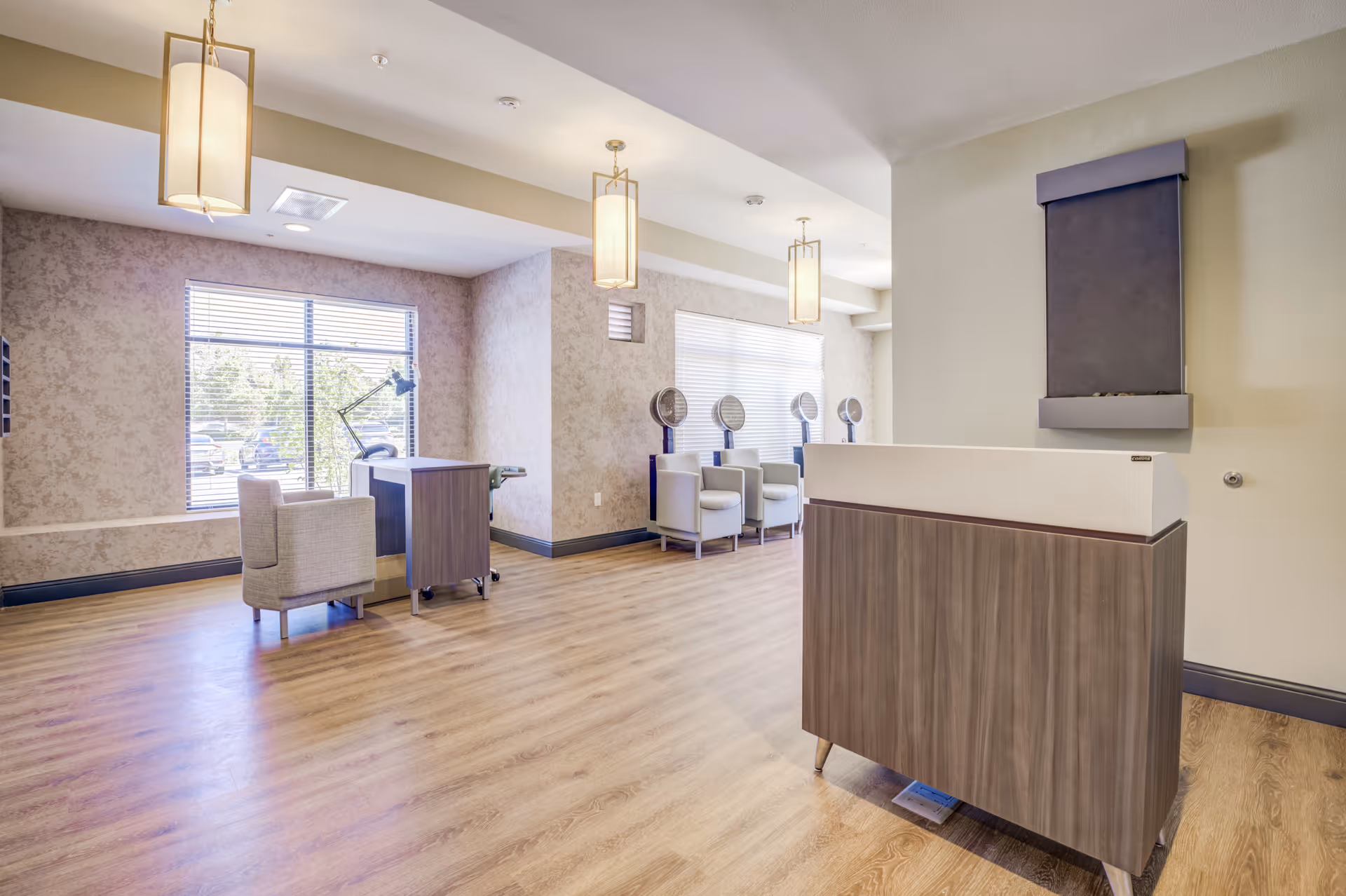 Interior view of a salon area in a senior living facility with wooden flooring, beige walls, and large windows letting in natural light. The room features a reception desk in the foreground, a manicure station with a chair and table near the window, and a row of salon chairs with hair dryers along the back wall. Modern pendant lights hang from the ceiling.