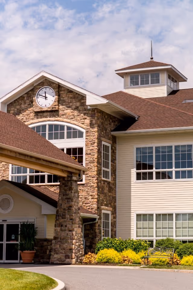 Exterior view of a senior living facility building with stone and beige siding, large windows, a clock on the stone facade, and a covered entrance with a stone pillar. There are green shrubs and yellow flowers near the entrance and a bench on the right side.