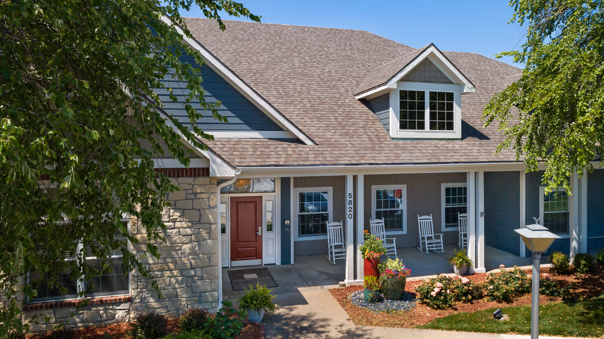 Front exterior view of a residential-style assisted living facility with a stone and blue siding facade, a red front door, white rocking chairs on the porch, and landscaped flower beds under a clear blue sky.