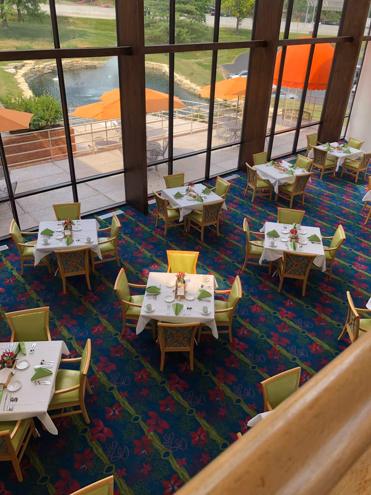 Interior view of a dining room with tables covered in white tablecloths, each set with green napkins, plates, cups, and silverware. The room has a colorful carpet with a floral pattern and large floor-to-ceiling windows showing an outdoor patio area with orange umbrellas and greenery.
