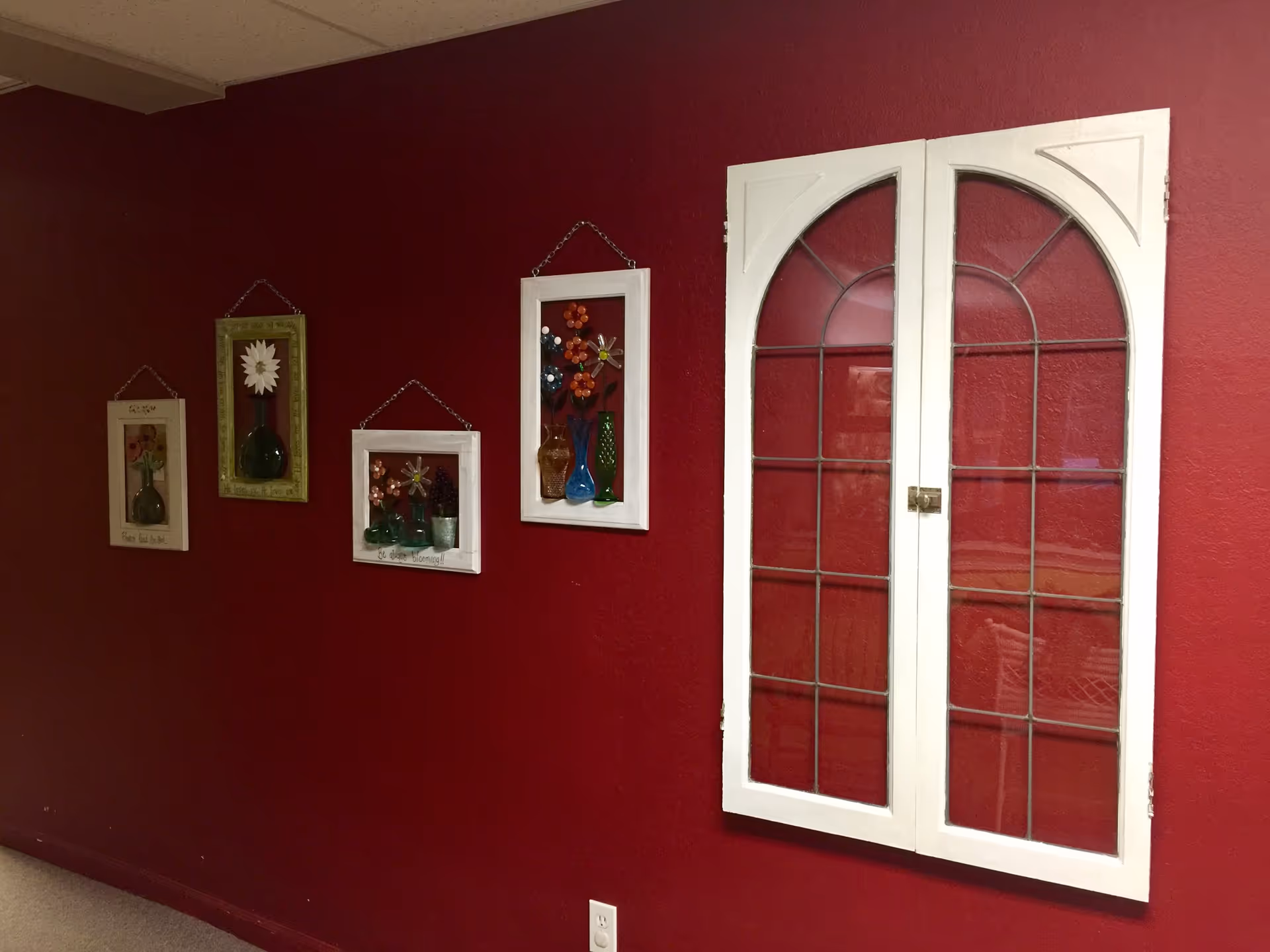 A deep red wall with four decorative framed flower art pieces hanging in a row. To the right, there is a white-framed window with an arched top and grid pattern. Below the window is an electrical outlet.