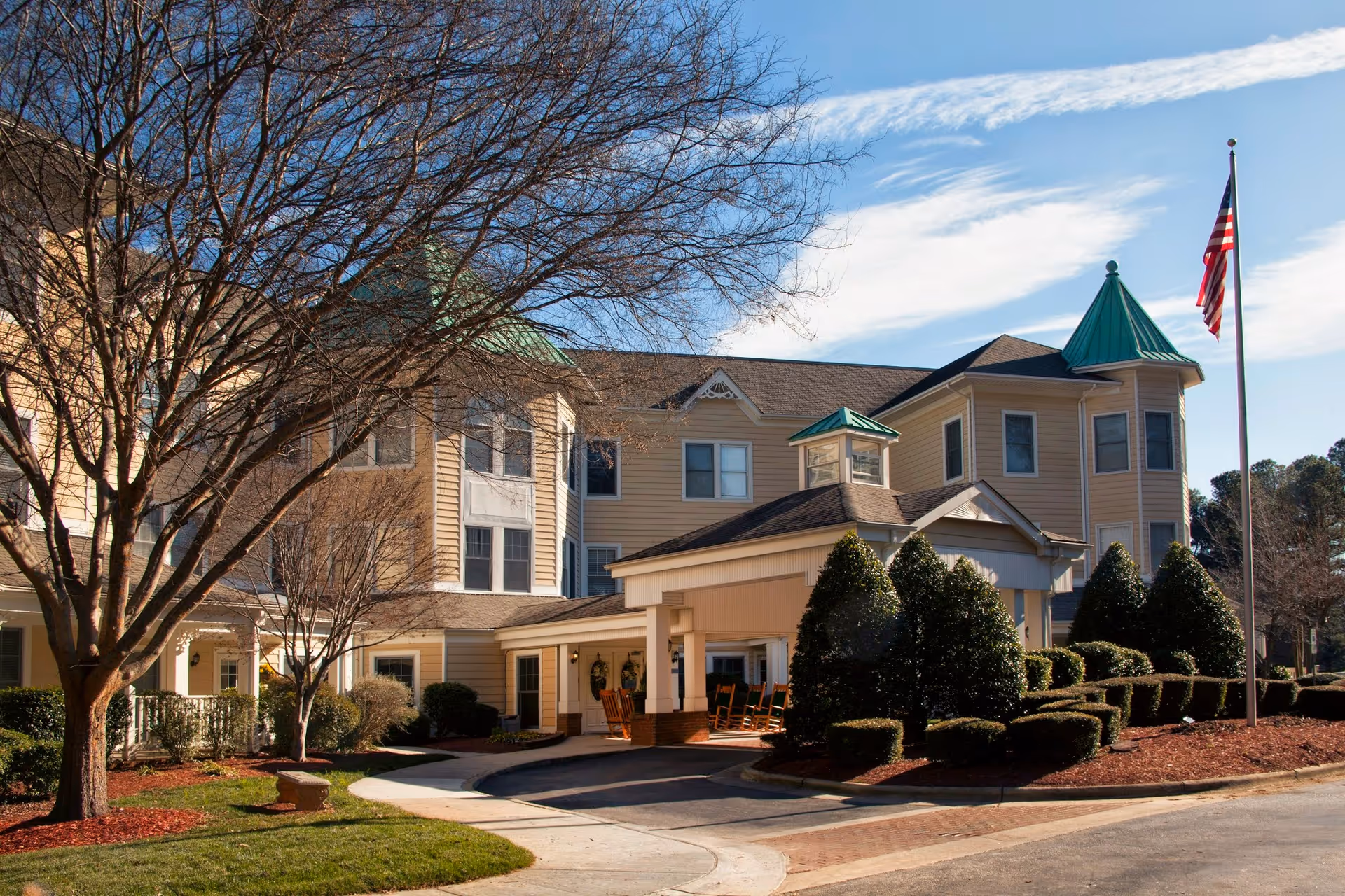 Exterior view of a senior living facility building with beige siding, green roof accents, and a covered entrance with rocking chairs. There are manicured bushes, a tree without leaves, and an American flag on a flagpole in front of the building under a partly cloudy sky.