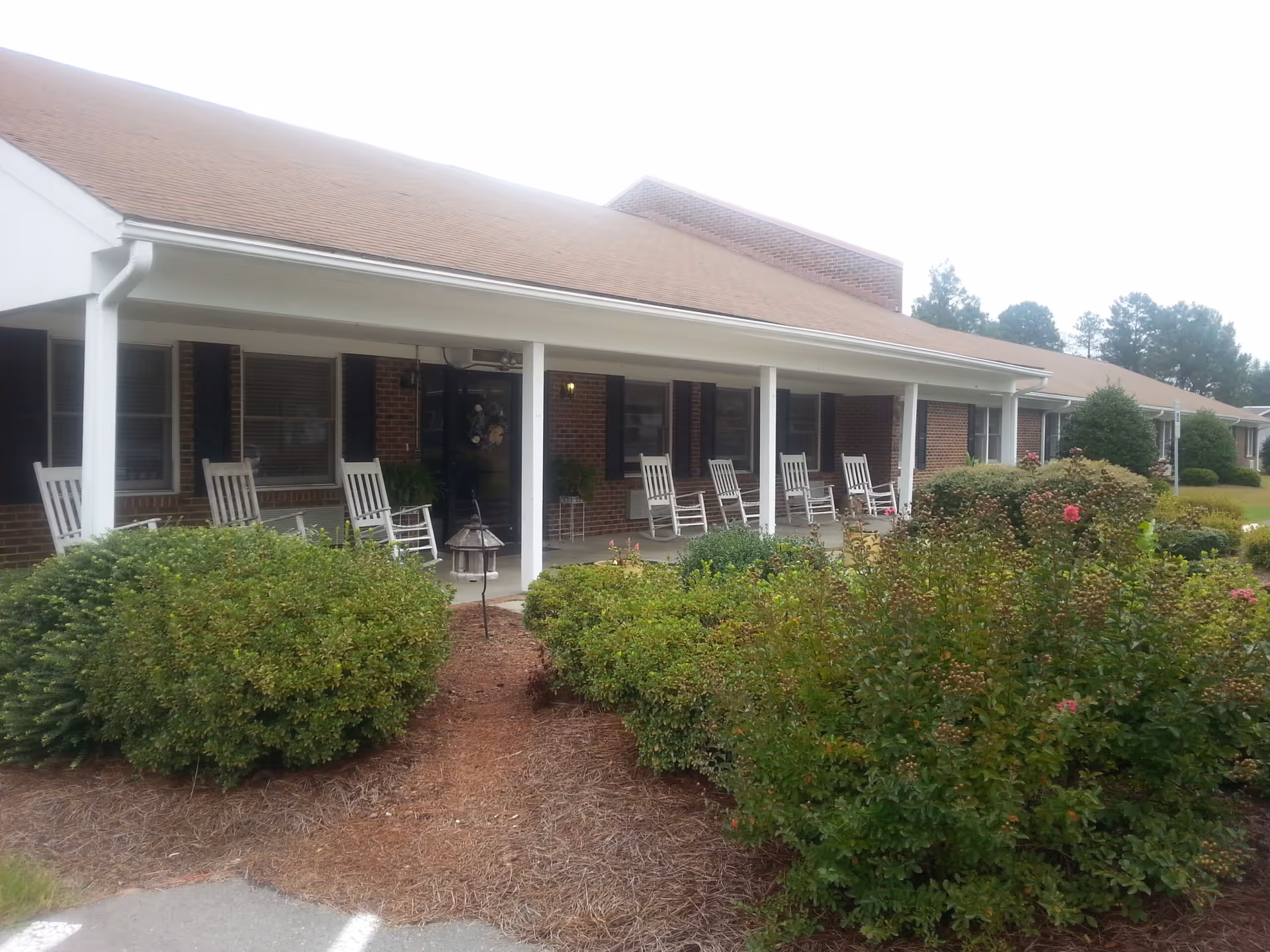 Long brick assisted living facility with a covered porch and white rocking chairs lining the walkway.
