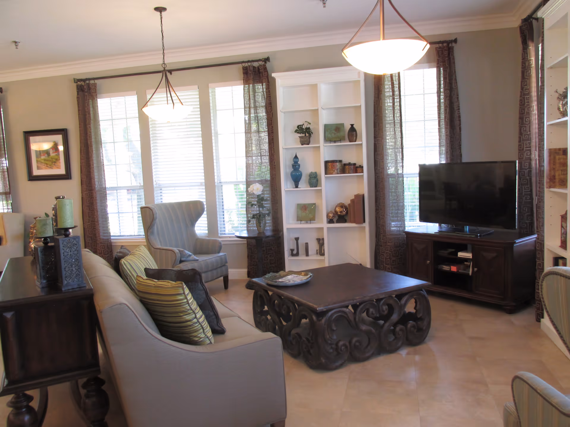 A well-lit living room with large windows covered by sheer curtains. The room features a beige sofa with striped pillows, a patterned armchair, a dark ornate coffee table, a wooden TV stand with a flat-screen television, and built-in white shelves decorated with vases and books. Two hanging pendant lights illuminate the space.