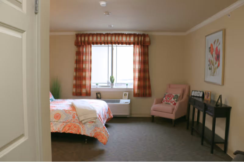 A cozy bedroom with a bed covered in a floral patterned bedspread, a pink armchair with a decorative pillow, a window with red plaid curtains, a small black console table with picture frames and a clock, and a framed floral artwork on the wall.