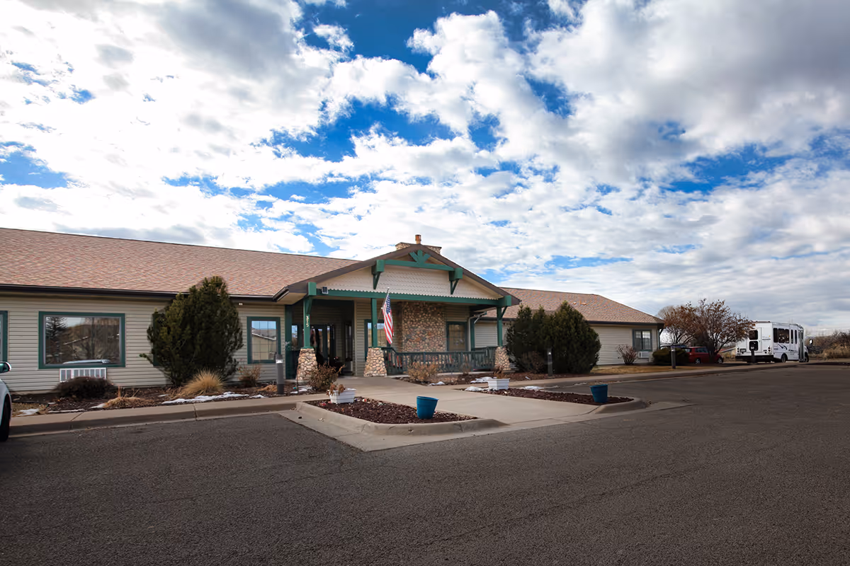 Exterior view of a single-story building with a peaked roof and a covered entrance supported by stone pillars. The building has beige siding with green trim and an American flag hanging near the entrance. There are shrubs and small trees around the building, a paved driveway, and a partly cloudy sky overhead.