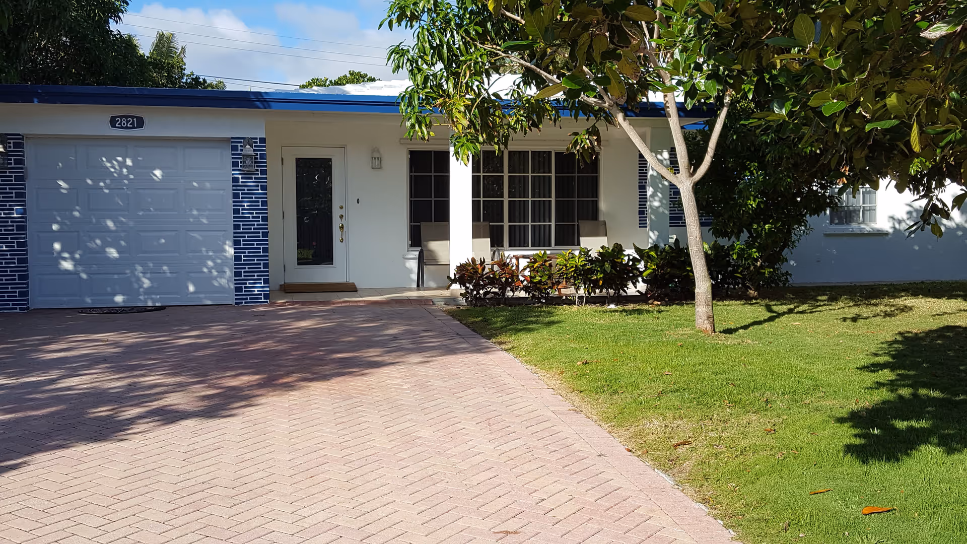 Front exterior of a single-story house with a garage, paved driveway, porch seating, and a grassy lawn with a tree.