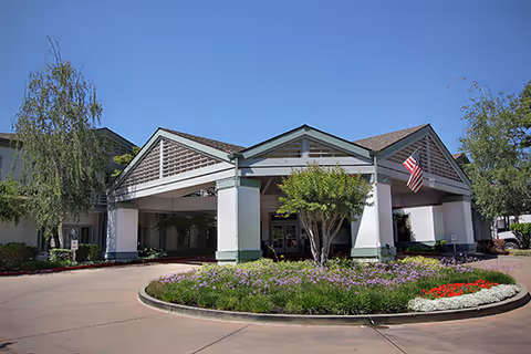 Front exterior view of Brookdale Diablo Lodge featuring a covered entrance with a triangular roof, an American flag, landscaped flower beds, and trees under a clear blue sky.