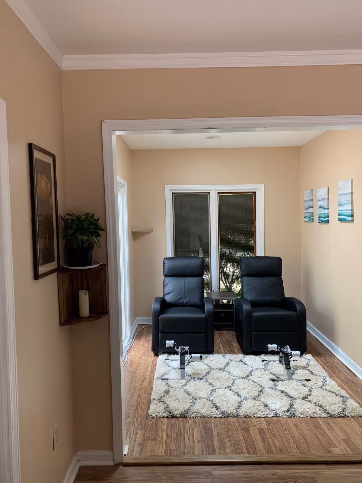 A small room with two black recliner chairs placed side by side on a patterned white and gray rug. Behind the chairs is a small table and a set of glass doors with blinds. The walls are painted beige, with three small blue and green abstract paintings on the right wall. To the left of the room entrance, there is a corner shelf holding a potted plant and a candle, and a framed flower picture hangs above it. The floor is wood laminate.