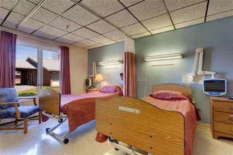 Two hospital-style beds in a shared patient room with teal walls and window view.