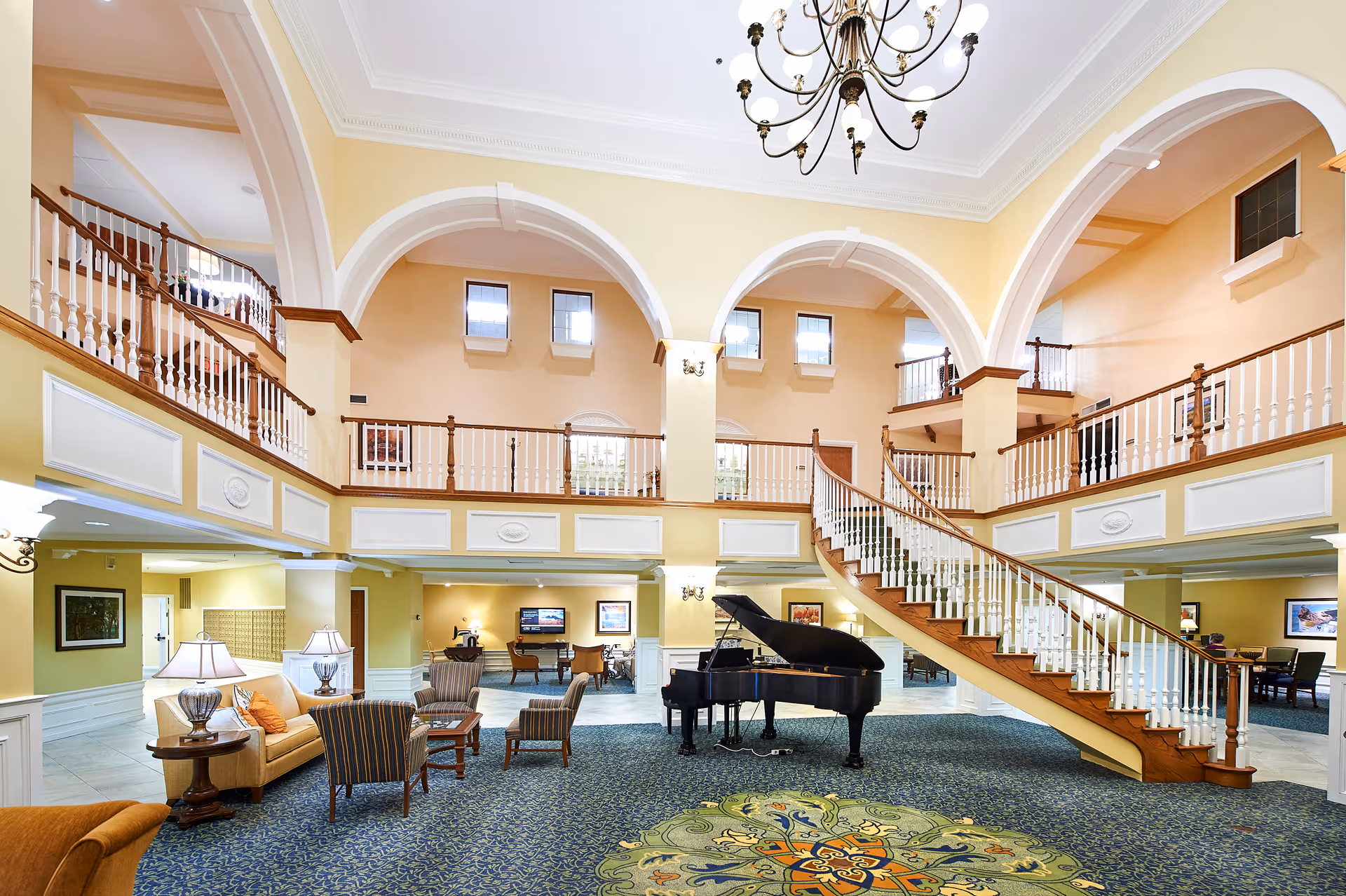 Spacious and elegant senior living facility common area with a grand piano in the center, surrounded by comfortable seating including armchairs and sofas. The room features high ceilings with large arches, a decorative chandelier, and a curved staircase leading to an upper balcony. The walls are painted in warm tones and the floor is covered with patterned carpet.