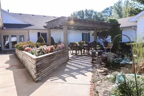 Sunlit outdoor courtyard with a wooden pergola, table and chairs, raised flower beds and a small pond beside a single-story building.