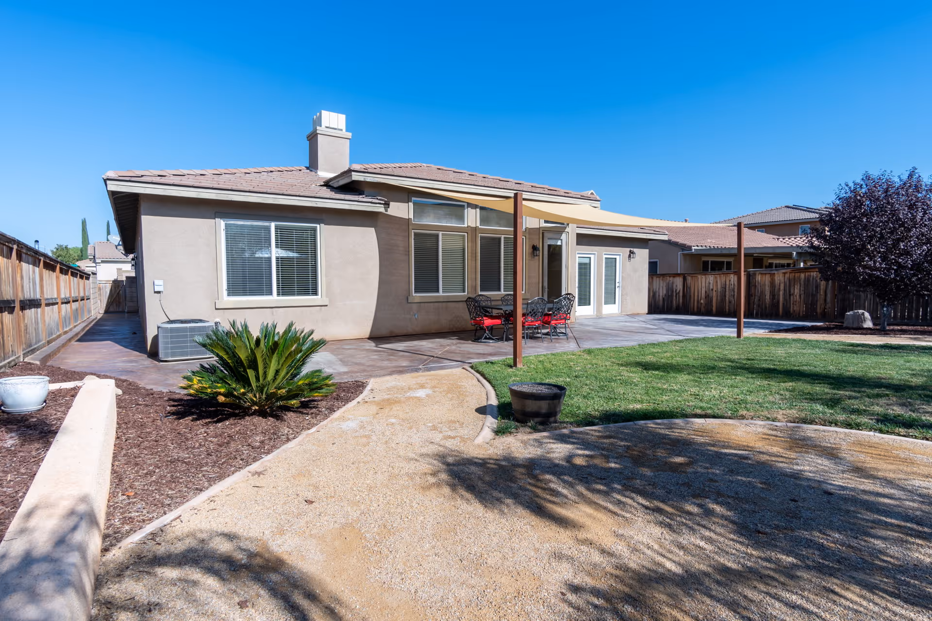 Backyard view of a single-story house with beige exterior walls and a tiled roof. The yard features a concrete patio with a table and chairs with red cushions under a beige canopy, a green lawn, a small tree, and a dirt pathway bordered by mulch and plants. A wooden fence encloses the yard.