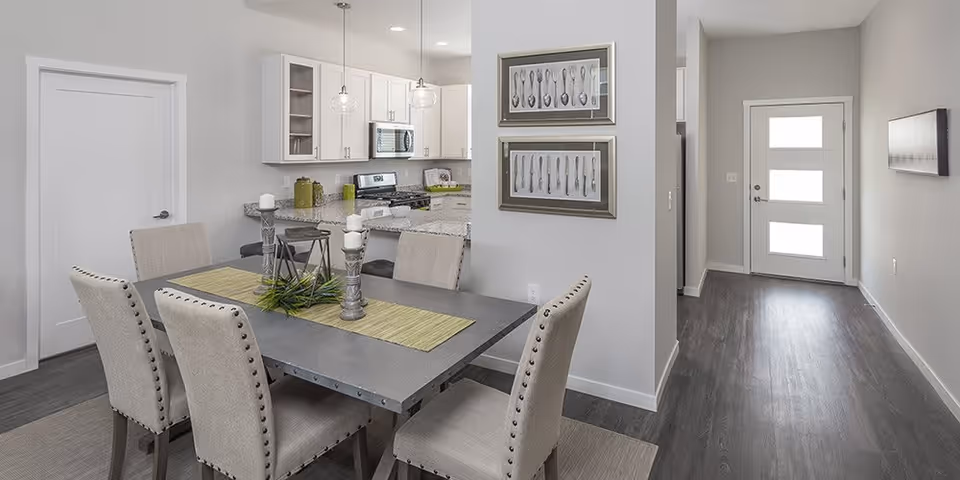 A modern dining area with a rectangular table set for six with beige upholstered chairs. The table has a green runner and decorative candles and greenery. In the background, there is a kitchen with white cabinets, a stove, microwave, and two pendant lights hanging above the counter. To the right, there is a hallway leading to a door with three frosted glass panels. The walls are light gray and the floor is dark wood.