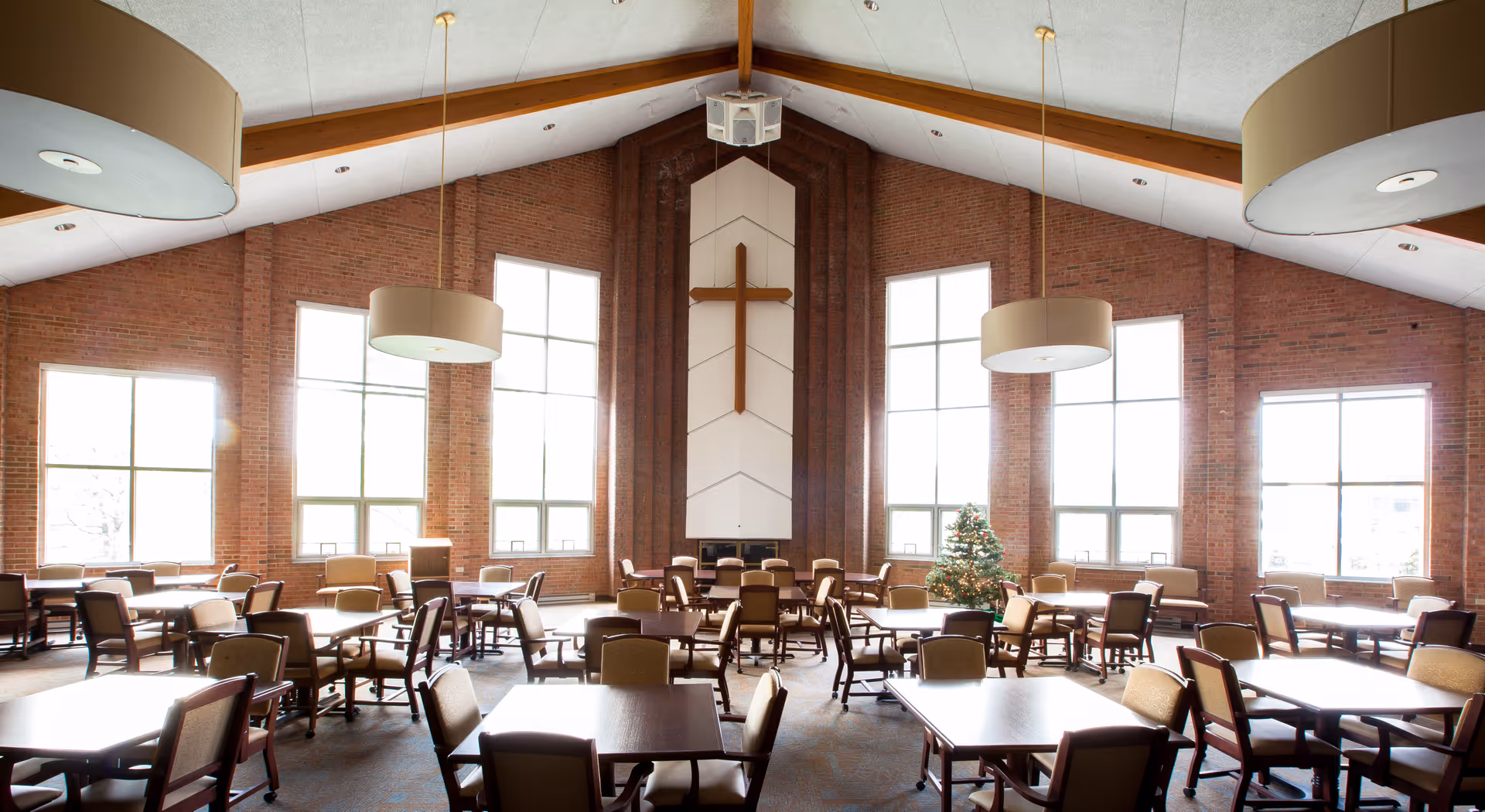 Spacious dining/activity room with many tables and chairs, high vaulted ceiling, large windows, a cross on the far wall and a small Christmas tree.