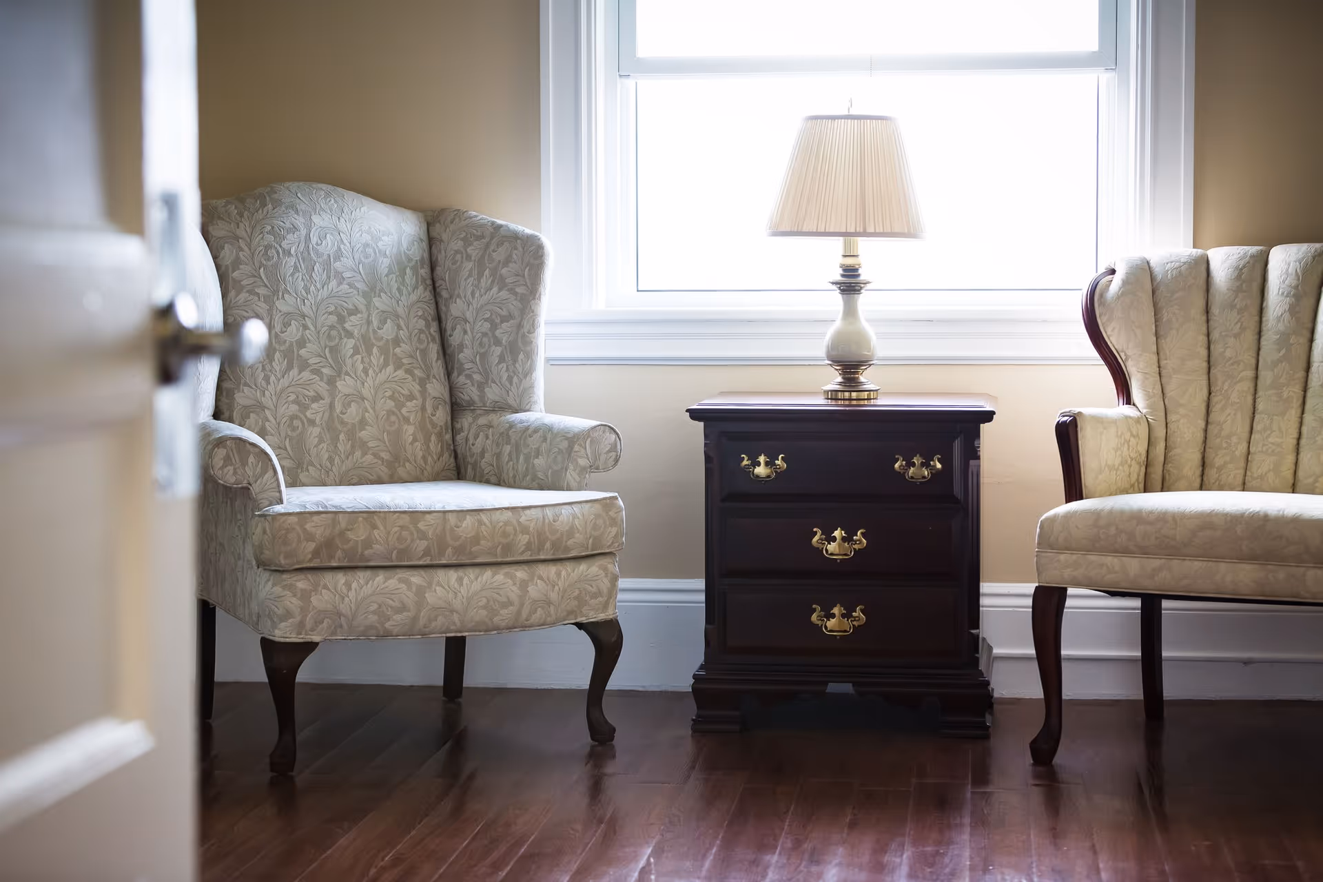 A cozy interior corner with two upholstered armchairs featuring a floral pattern, separated by a dark wooden side table with three drawers and brass handles. A table lamp with a beige shade sits on the side table in front of a bright window.