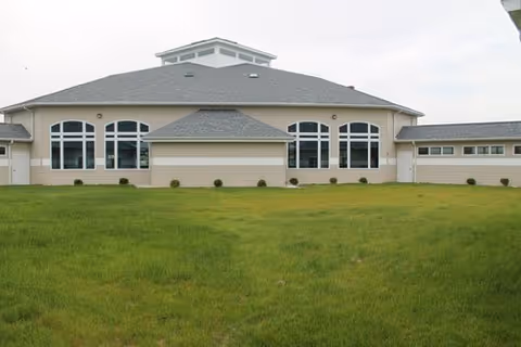 Exterior view of a single-story building with multiple large windows and a gray roof, surrounded by a green lawn under a cloudy sky.