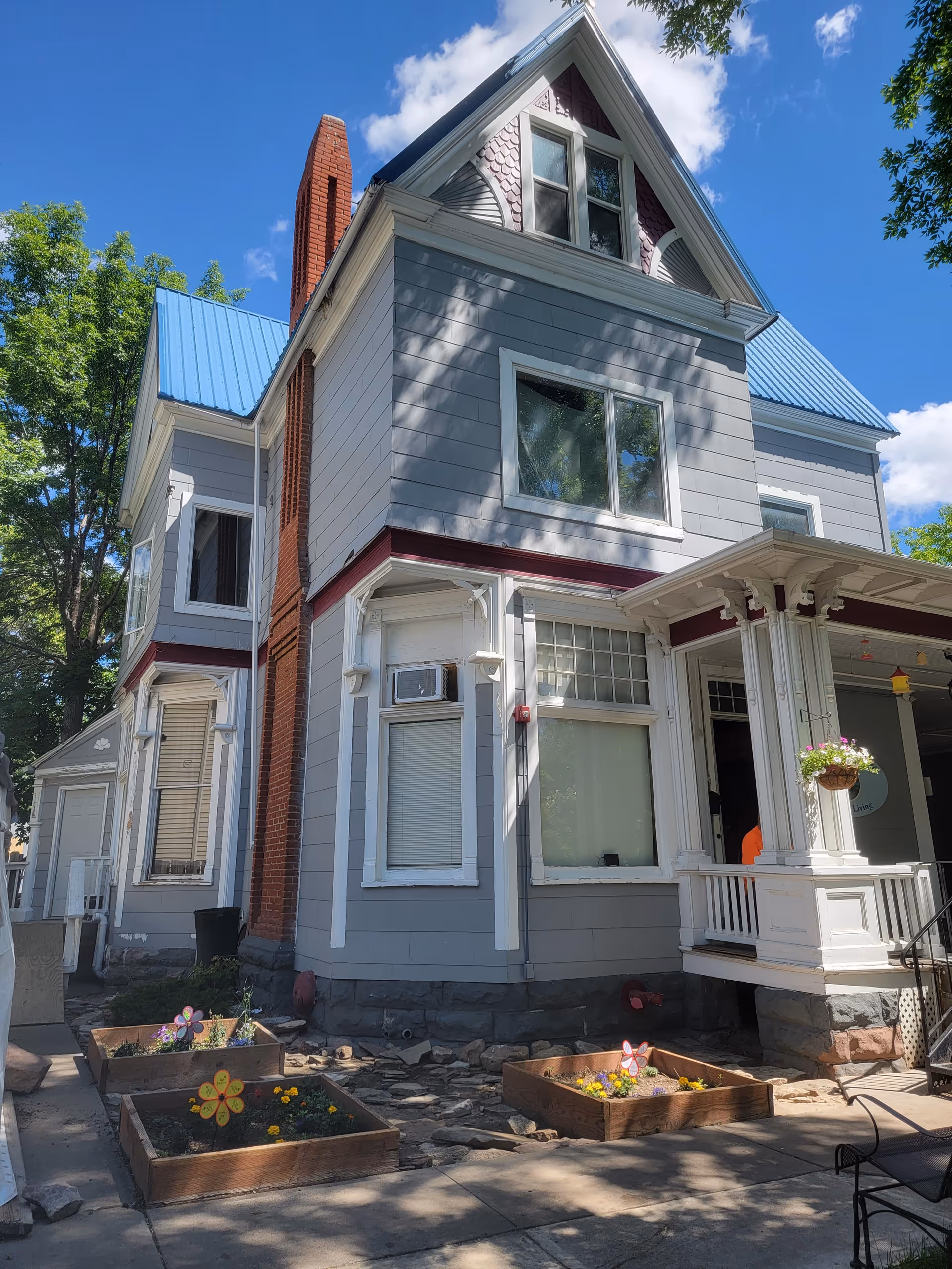 Exterior view of a two-story house with gray siding, white trim, and a blue metal roof under a clear blue sky. The house has a small porch with decorative columns and hanging flower baskets. There are three raised garden beds with flowers in front of the house and a brick chimney on the side.