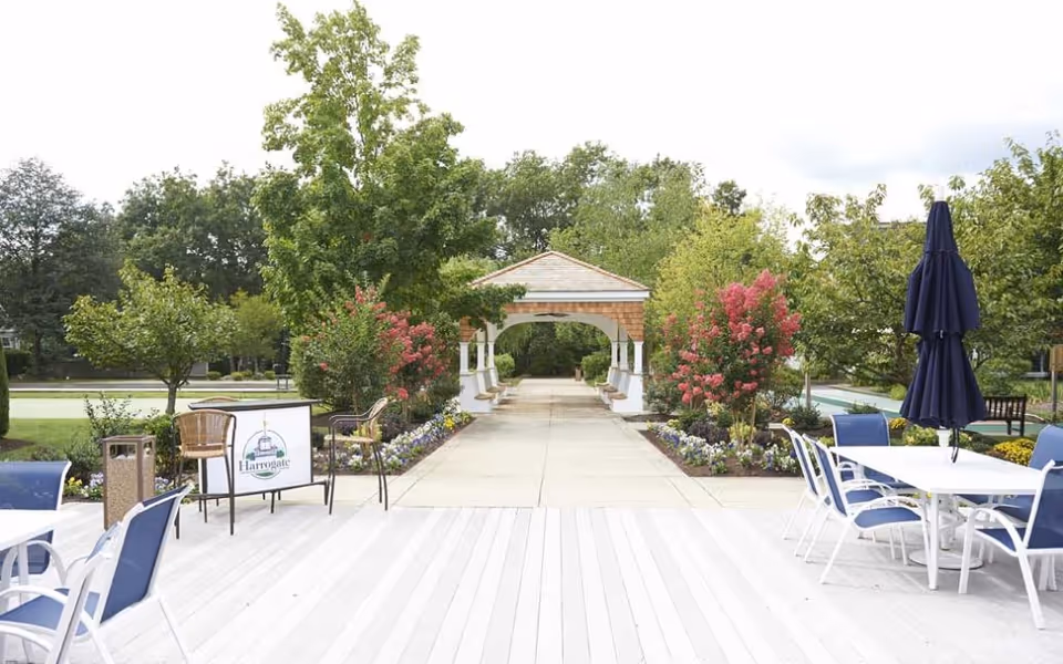 Outdoor patio area at Harrogate Retirement Community with white tables and blue chairs, a walkway lined with flowers and trees leading to a small pavilion with benches, and a sign displaying the Harrogate logo.