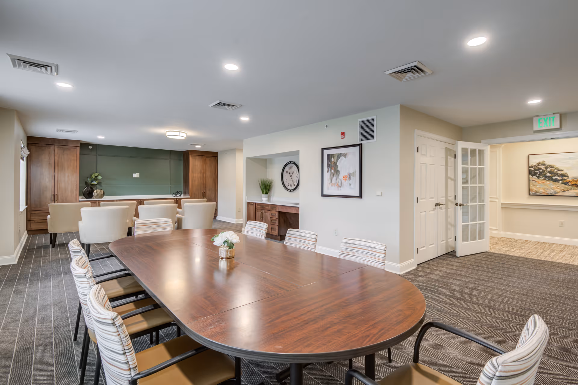 A well-lit meeting or dining room with a large wooden oval table surrounded by chairs with striped cushions. In the background, there is a seating area with beige upholstered chairs and wooden cabinets against a green accent wall. The room has carpeted floors, recessed ceiling lights, a wall clock, framed artwork, and a door leading to another room with a painting on the wall.