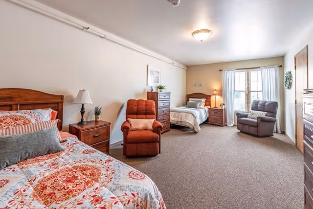 Spacious senior memory care bedroom with two twin beds, recliners, dressers and a window letting in natural light.