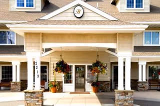 Front exterior view of a senior living facility with a covered entrance supported by white columns and stone bases, hanging flower baskets, and windows on the upper and lower floors.