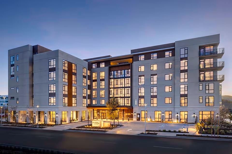 Exterior view of a modern multi-story senior living facility building at dusk with lights glowing from the windows and a well-lit entrance area.