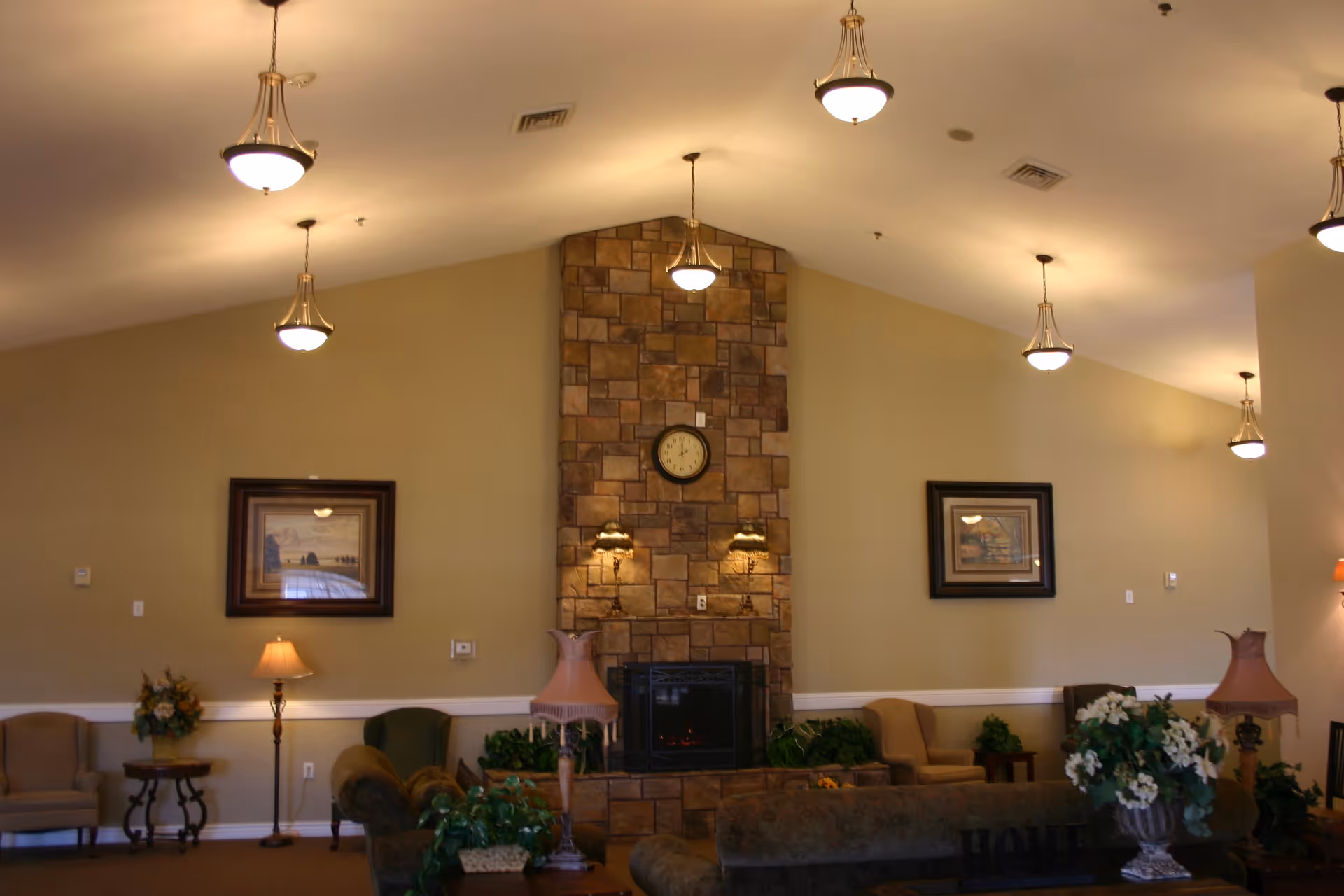 A spacious living room area with a high vaulted ceiling featuring multiple hanging light fixtures. The room has a central stone fireplace with a clock above it, flanked by two wall lamps. There are several armchairs and sofas arranged around the room, along with side tables, floor lamps, framed artwork on the walls, and decorative plants.