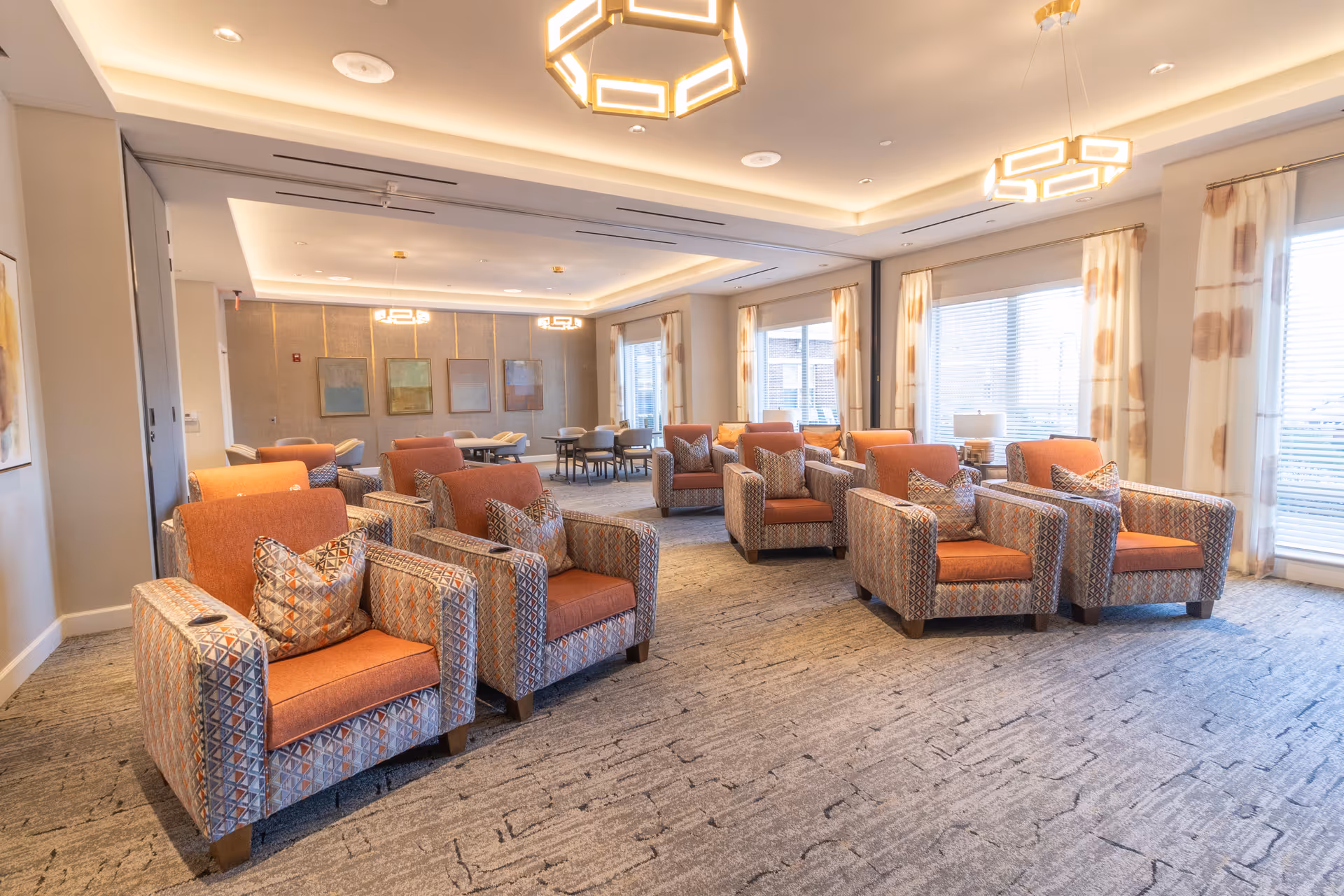 A spacious and well-lit living room area in a senior living facility with multiple patterned armchairs featuring orange cushions arranged in rows. The room has large windows with white and orange curtains, modern geometric ceiling lights, and a carpeted floor. In the background, there are tables and chairs along the far wall with framed artwork hanging above them.