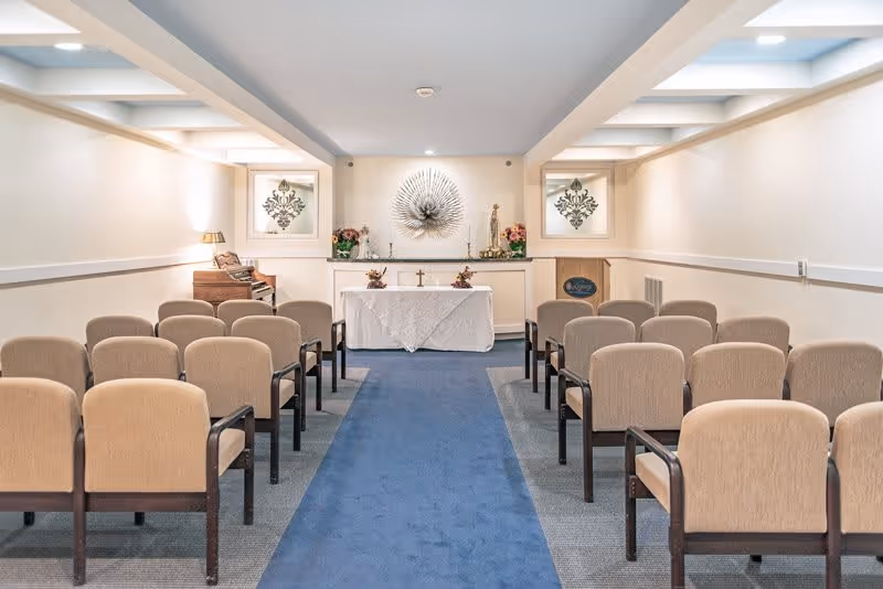 Interior view of a small chapel or meditation room with rows of beige cushioned chairs arranged on either side of a blue carpeted aisle leading to an altar covered with a white cloth. The altar has decorative items including flowers and a cross, with a large metallic sunburst wall decoration above it. The room has soft lighting and neutral-colored walls.