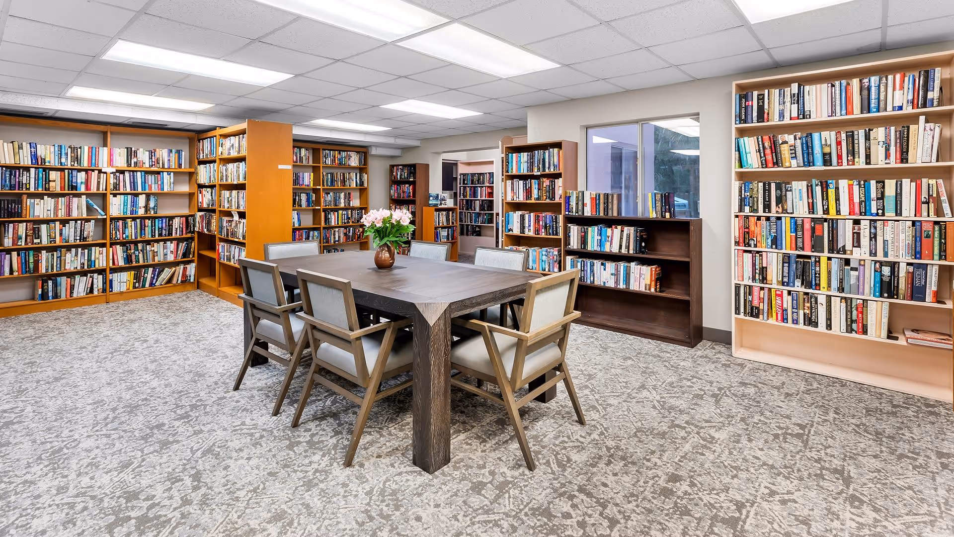 A well-lit library room with multiple wooden bookshelves filled with books. In the center, there is a dark wooden table surrounded by six cushioned chairs. A vase with pink flowers is placed on the table. The room has a patterned carpet and a window on the right wall.