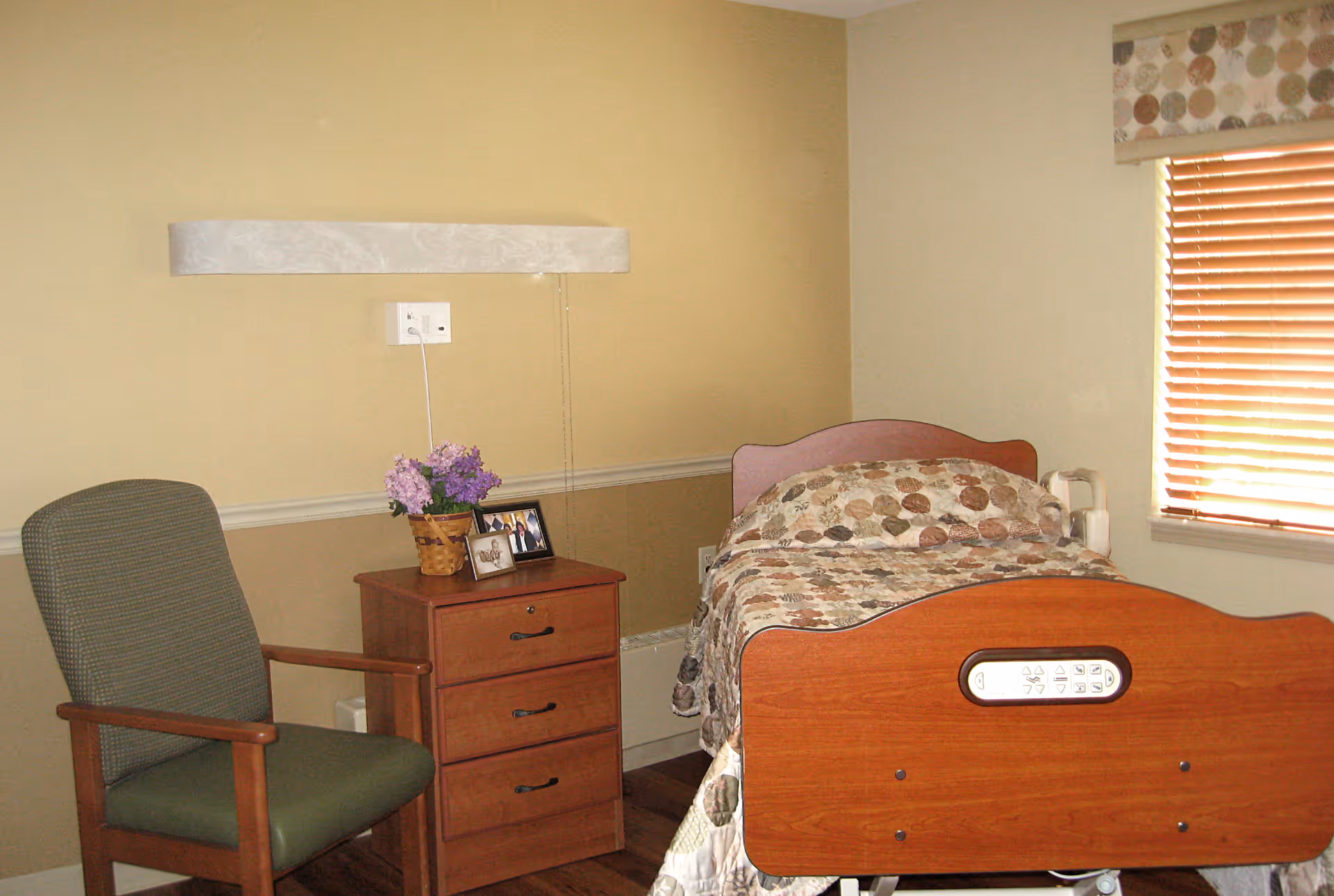 A small bedroom with a single hospital-style bed featuring a wooden headboard and footboard. The bed is covered with a patterned quilt. Next to the bed is a wooden nightstand with three drawers, a basket of purple flowers, and two framed photos. To the left of the nightstand is a green cushioned chair with wooden armrests. A window with wooden blinds and a valance is on the right wall, allowing natural light into the room. The walls are painted beige with a white chair rail molding.