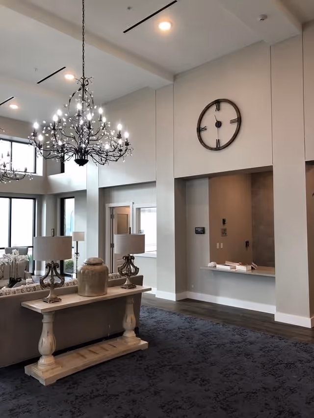 Open, modern lounge area with chandeliers, a large wall clock, a sofa table topped with lamps and a decorative urn, and a reception counter.