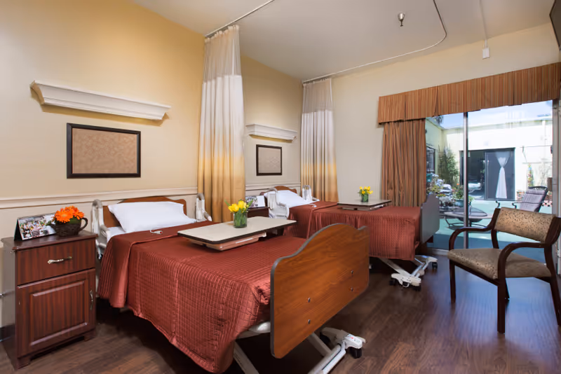 A shared bedroom in a senior living facility with two hospital-style beds covered in brown quilts. Each bed has a white pillow and a small overbed table with a vase of yellow flowers. There is a wooden nightstand with a photo frame and a flower pot on the left side. The room has beige walls, framed artwork, and large sliding glass doors leading to an outdoor patio area. A cushioned chair is positioned near the door.