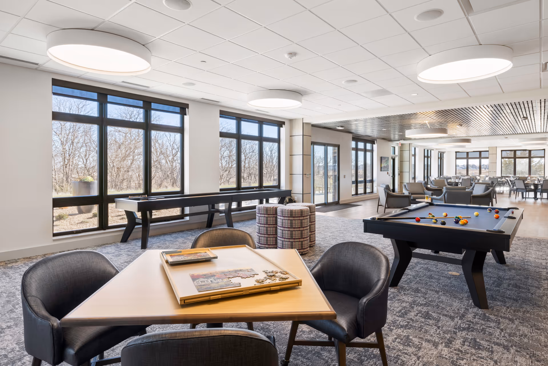 Bright senior living common room with large windows, seating areas, a pool table and a game table.